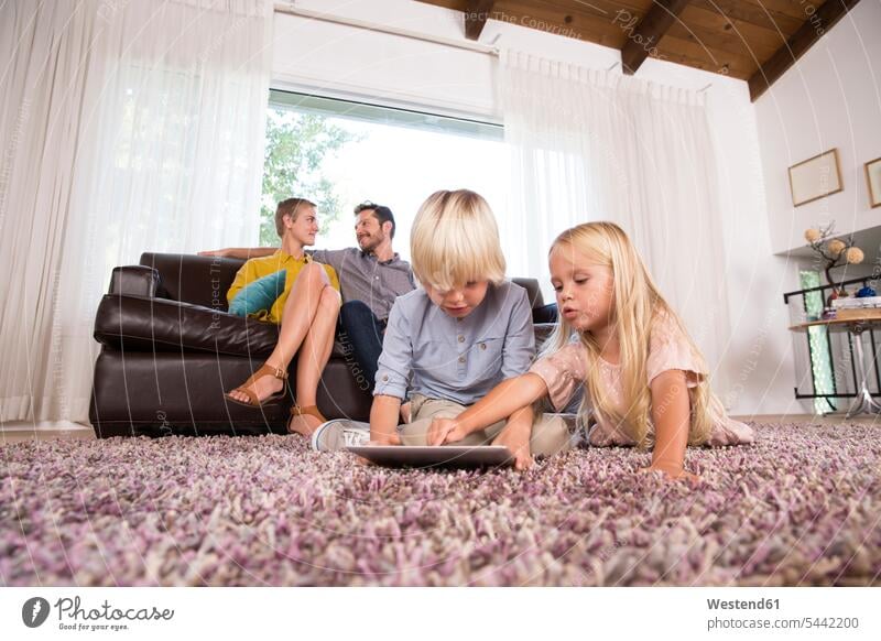 Bruder und Schwester benutzen Tablette auf Teppich im Wohnzimmer mit den Eltern im Hintergrund Brüder Schwestern Teppiche Tablet Computer Tablet-PC Tablet PC
