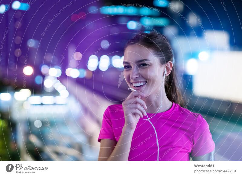 Lächelnde junge Frau im rosa Sporthemd, die nachts in der Stadt Musik hört junge Frauen telefonieren anrufen Anruf telephonieren sprechen reden Musik hören