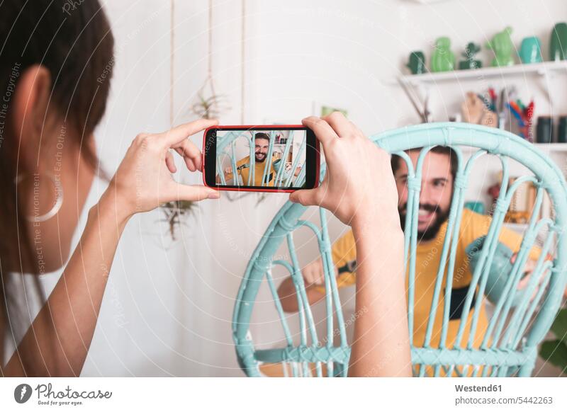 Junge Frau fotografiert bärtigen Mann, der zu Hause einen Korbsessel bemalt Handy Mobiltelefon Handies Handys Mobiltelefone fotografieren Paar Pärchen Paare