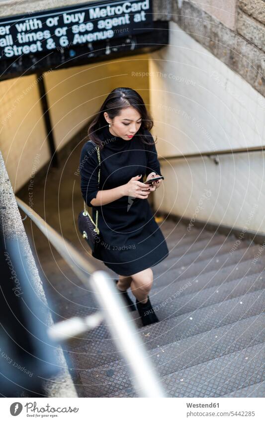 USA, New York City, Manhattan, junge Frau in Schwarz gekleidet, geht die Treppe hinauf und schaut auf ihr Handy weiblich Frauen Smartphone iPhone Smartphones
