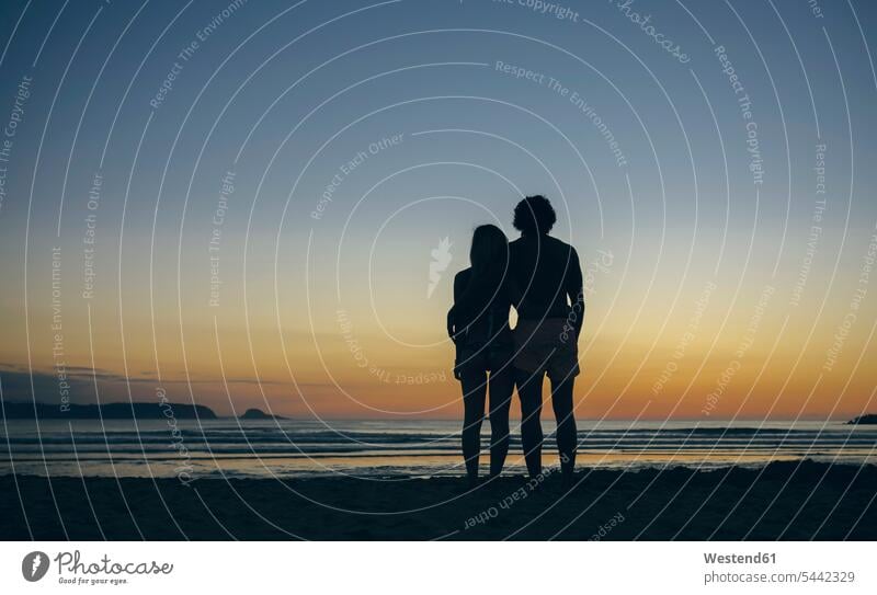 Junges Paar beobachtet den Sonnenuntergang am Strand Pärchen Paare Partnerschaft Beach Straende Strände Beaches Mensch Menschen Leute People Personen Urlaub