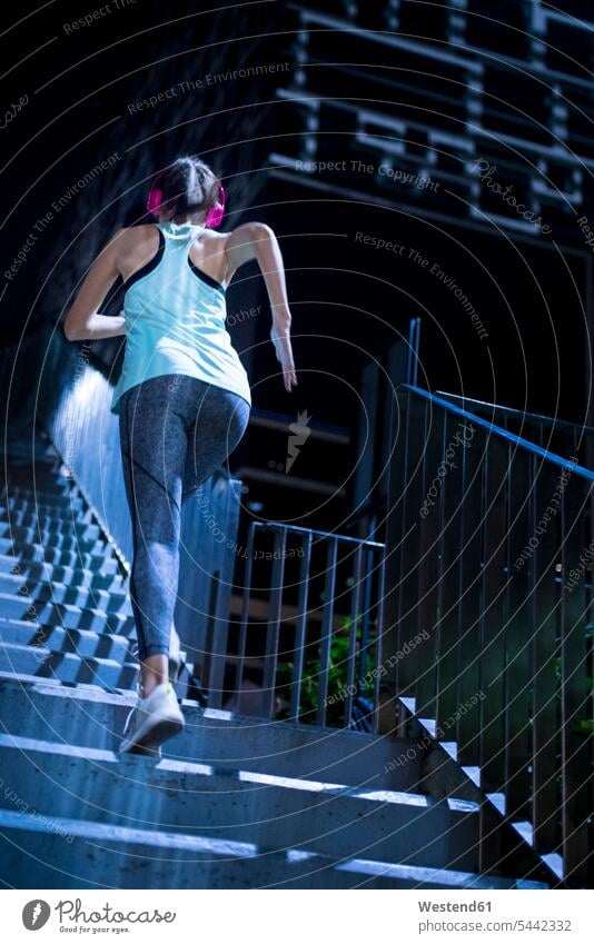 Junge Frau mit rosa Kopfhörern, die nachts in moderner städtischer Umgebung die Treppe hinaufläuft trainieren laufen rennen Joggerin Joggerinnen fit aktiv