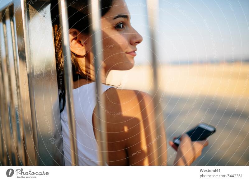 Lächelnde junge Frau lehnt bei Sonnenuntergang am Geländer weiblich Frauen Erwachsener erwachsen Mensch Menschen Leute People Personen Abend abends Smartphone