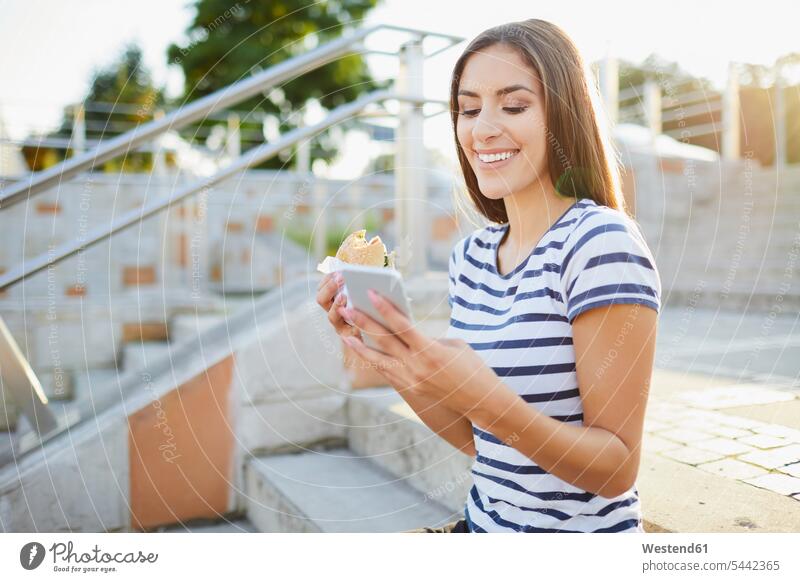 Junge Frau sitzt auf der Treppe, isst Bagel und benutzt Smartphone weiblich Frauen lächeln Handy Mobiltelefon Handies Handys Mobiltelefone essen essend