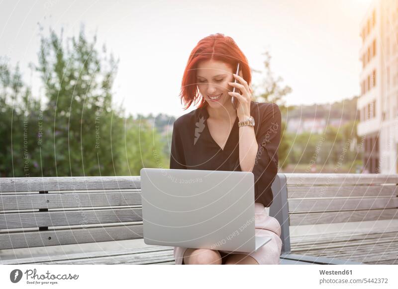 Junge Frau sitzt mit ihrem Laptop auf der Bank und telefoniert weiblich Frauen gut gekleidet Smart Casual gut angezogen Geschäftskleidung jung Smartphone iPhone