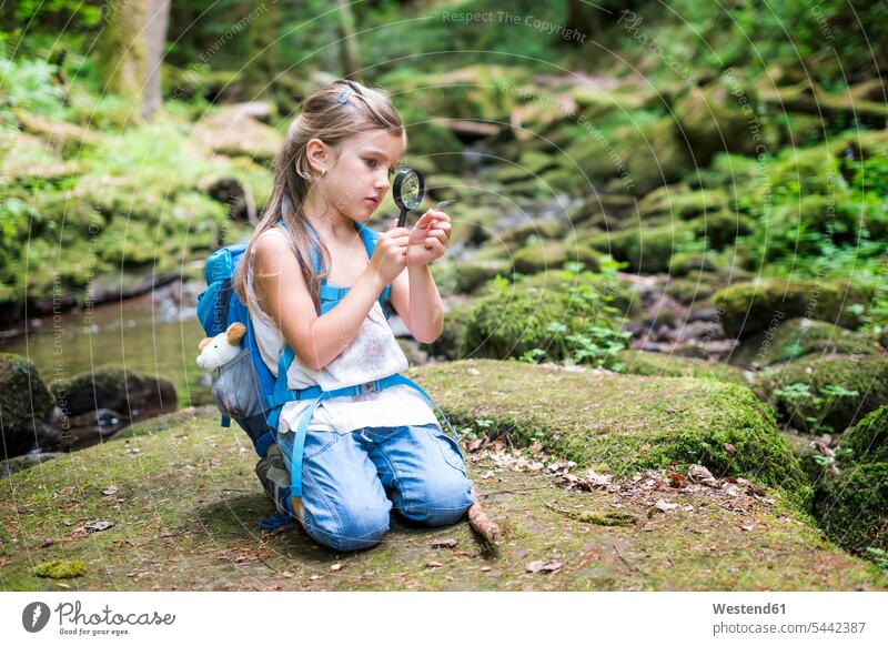 Kleines Mädchen mit Lupe, das auf einem Felsen im Wald kauert und eine Feder beobachtet betrachten betrachtend weiblich Leselupen Lupen schauen sehend Kind