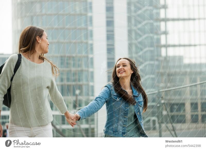 Zwei Freundinnen, die in der Stadt spazieren gehen und sich an den Händen halten gehend geht lachen fröhlich Fröhlichkeit Frohsinn Heiterkeit glücklich Glück