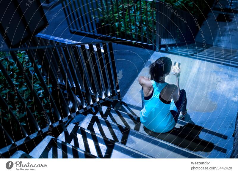 Junge Frau sitzt auf einer Treppe und überprüft nachts ihr Smartphone in einer modernen städtischen Umgebung iPhone Smartphones trainieren Joggerin Joggerinnen