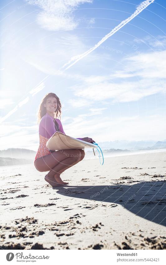 Lächelnde Frau mit Surfbrett kauert am Strand Beach Straende Strände Beaches lächeln Surferin Wellenreiterinnen Surferinnen hocken kauernd hockend Surfbretter