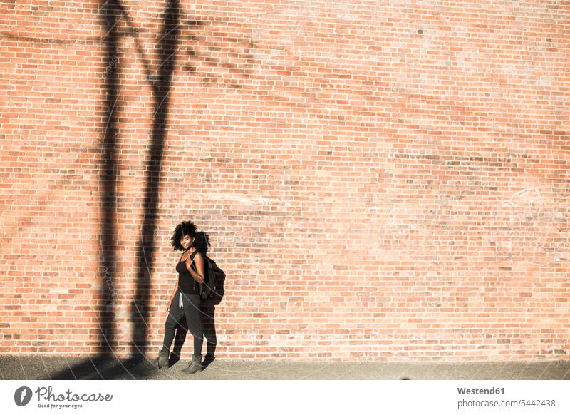 Frau mit Rucksack vor einer Ziegelmauer mit Schatten eines Strommastes stehen stehend steht weiblich Frauen Backsteinwand Backsteinmauern Erwachsener erwachsen