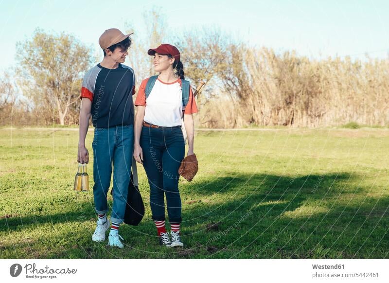 Lächelndes junges Paar mit Baseballausrüstung geht im Park spazieren Baseballspiel lächeln Pärchen Paare Partnerschaft Baseballspieler Baseballer Sport Mensch