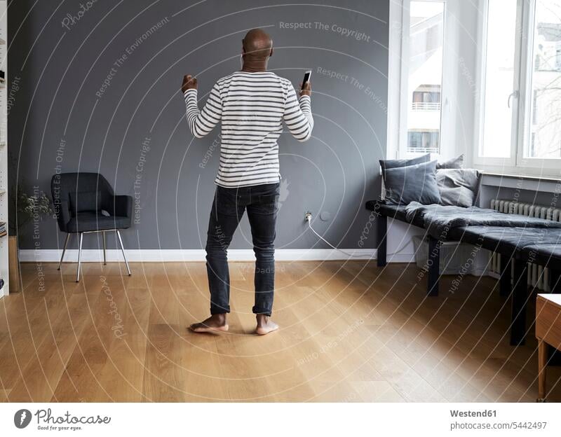 Reifer Mann, der allein zu Hause tanzt und ein Smartphone in der Hand hält Zuhause daheim barfuß nackte Füße nackter Fuss barfuessig barfüssig nackter Fuß
