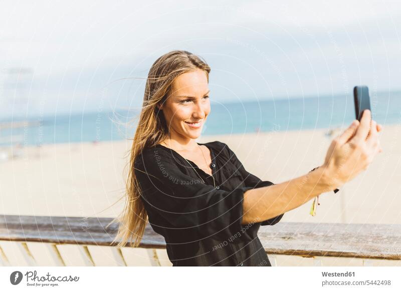 Lächelnde junge Frau macht ein Selfie am Strand Selfies lächeln weiblich Frauen glücklich Glück glücklich sein glücklichsein Erwachsener erwachsen Mensch
