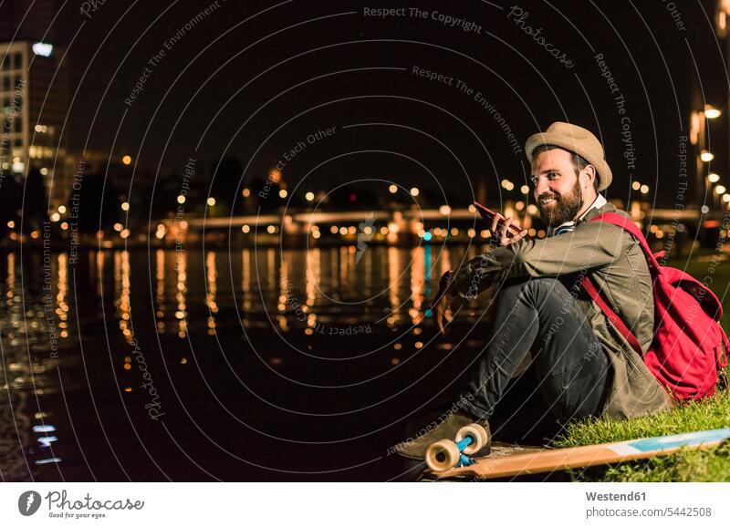 Lächelnder junger Mann mit Handy und Skateboard sitzt nachts am städtischen Flussufer lächeln Nacht Männer männlich Erwachsener erwachsen Mensch Menschen Leute