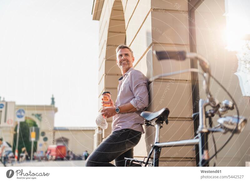 Lächelnder Mann mit Fahrrad und Kaffee zum Mitnehmen in der Stadt staedtisch städtisch lächeln Bikes Fahrräder Räder Rad Männer männlich Außenaufnahme draußen