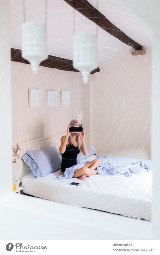 Junge Frau sitzt im Bett und trägt eine VR-Brille sitzen sitzend Virtuelle Realität Virtuelle Realitaet Brillen weiblich Frauen Betten Erwachsener erwachsen