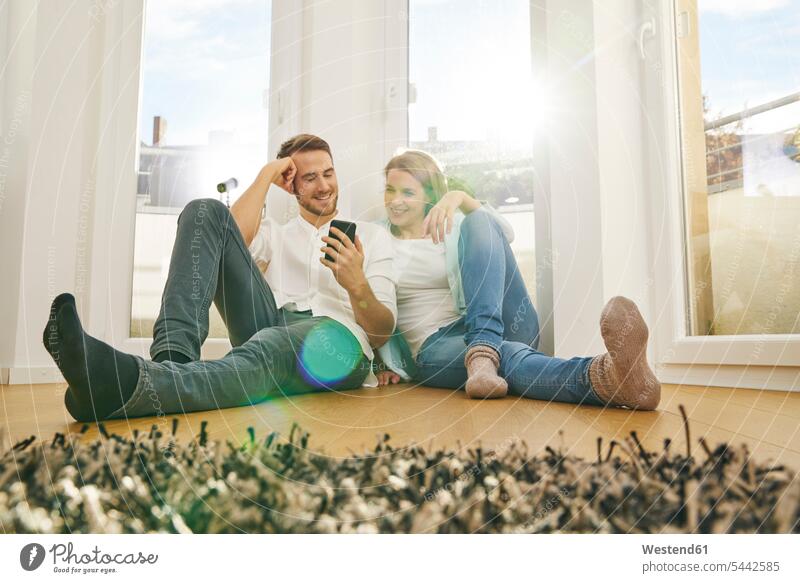 Lächelndes Paar sitzt auf dem Boden und schaut auf sein Handy lächeln Mobiltelefon Handies Handys Mobiltelefone Pärchen Paare Partnerschaft Telefon telefonieren