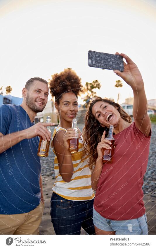 Drei Freunde mit Bierflaschen machen ein Selfie am Strand Beach Straende Strände Beaches Selfies Freundschaft Kameradschaft fotografieren Smartphone iPhone