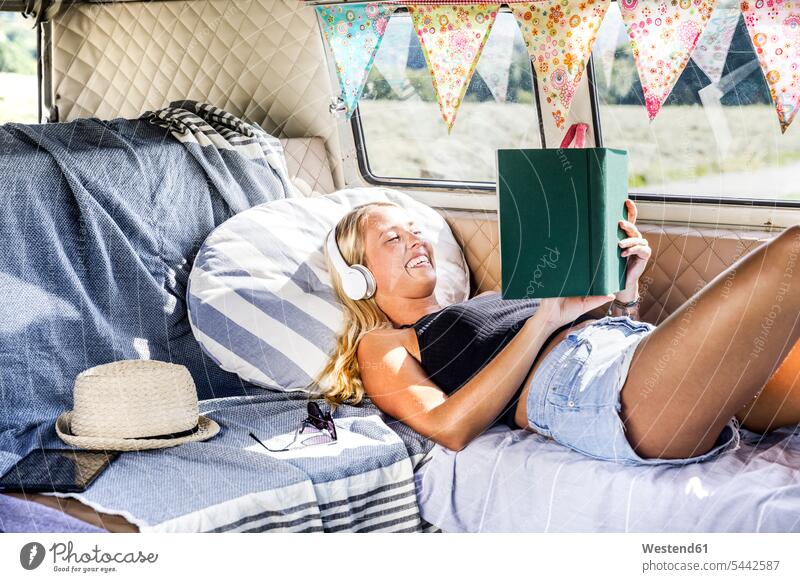 Glückliche Frau mit Kopfhörern liegt in einem Lieferwagen und liest ein Buch Bücher lachen weiblich Frauen lesen Lektüre Kleinbus Kleinbusse positiv Emotion