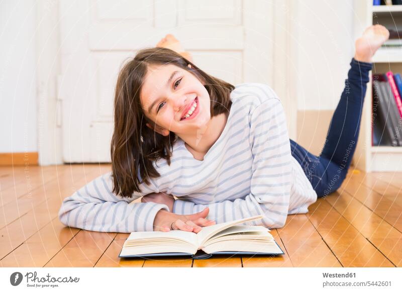 Porträt eines glücklichen Mädchens auf dem Boden liegend mit Buch Fußboden Portrait Glück Kind Mensch gute Laune Holzboden Bücherregal Freude Freizeitkleidung