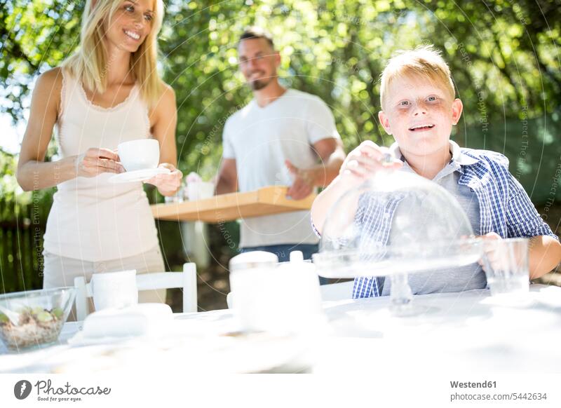 Lächelnder Junge mit seinen Eltern am Gartentisch Buben Knabe Jungen Knaben männlich Familie Familien Sohn Söhne lächeln Tisch Tische Kind Kinder Kids Mensch