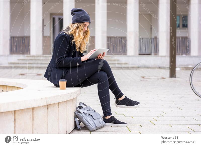 Junge Frau mit Kaffee zum Sitzen an der Wand mit Tablette benutzen Coffee to go Mauer sitzen Erwachsener Mensch Computer Getränk zum mitnehmen Food and Drink
