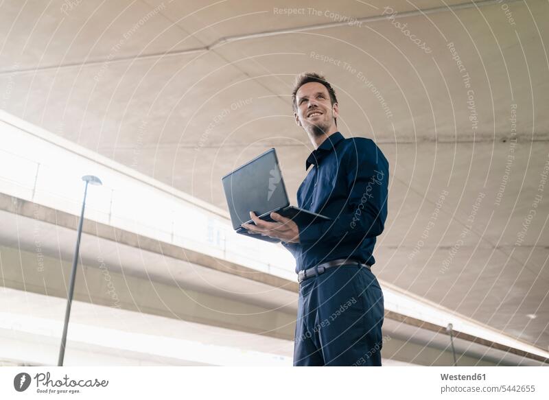 Zuversichtlicher Geschäftsmann steht mit Laptop an der Unterführung Notebook Laptops Notebooks lächeln Businessmann Businessmänner Geschäftsmänner Computer