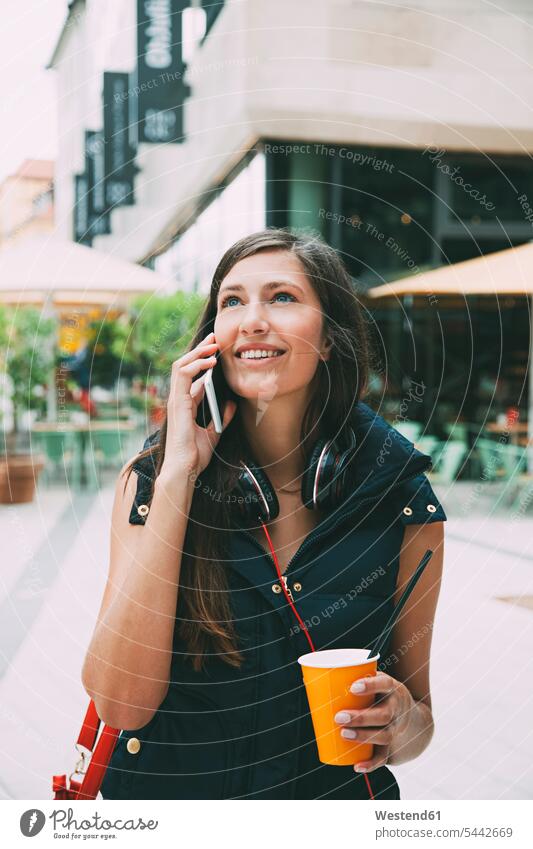 Porträt einer lächelnden jungen Frau am Handy mit Getränk zum Mitnehmen in der Stadt weiblich Frauen Mobiltelefon Handies Handys Mobiltelefone telefonieren