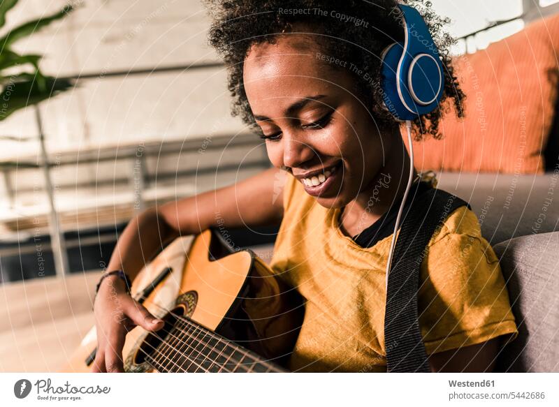 Lächelnde junge Frau zu Hause mit Kopfhörern, die Gitarre spielt lächeln weiblich Frauen Gitarren Kopfhoerer Erwachsener erwachsen Mensch Menschen Leute People