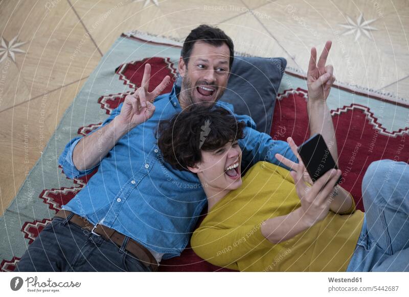 Paar liegt zusammen auf dem Teppich und macht ein Selfie mit Smartphone Selfies Pärchen Paare Partnerschaft Mensch Menschen Leute People Personen schreien rufen