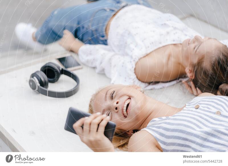 Zwei glückliche junge Frauen mit Handy und Kopfhörern in einem Skatepark Freundinnen Mobiltelefon Handies Handys Mobiltelefone Skateboardpark Skateboard-Park