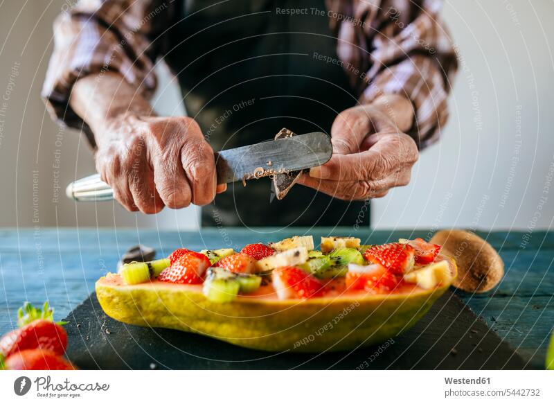 Nahaufnahme von Männerhänden, die Schokolade über eine halbe Papaya mit Banane, Kiwi und Erdbeeren schneiden Papayas Carica papaya Mann männlich Zubereitung