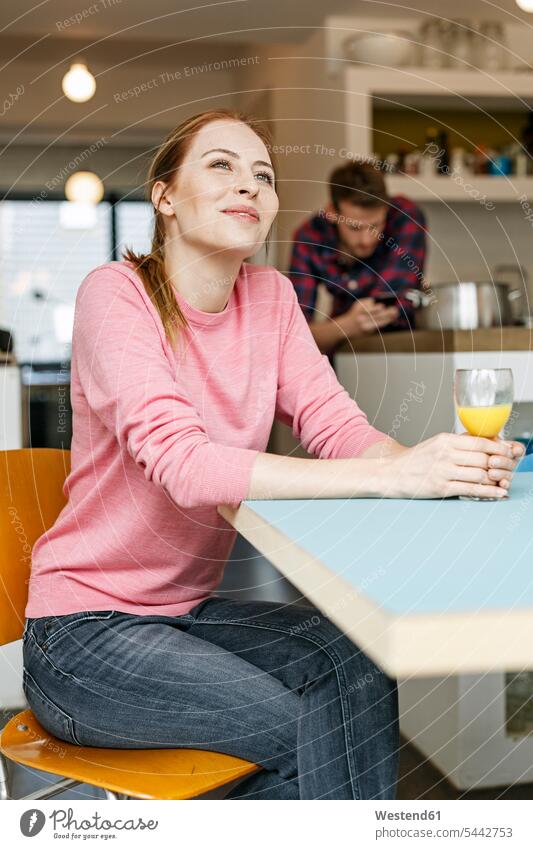 Lächelnde junge Frau mit einem Glas Orangensaft in der heimischen Küche mit einem Mann im Hintergrund Zuhause zu Hause daheim weiblich Frauen Küchen lächeln