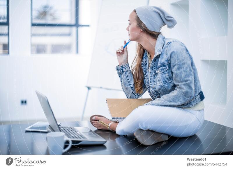 Junge Frau, die im Büro arbeitet und im Schneidersitz mit ihrem Laptop auf dem Schreibtisch sitzt Arbeitstisch Schreibtische sitzen sitzend Notebook Laptops
