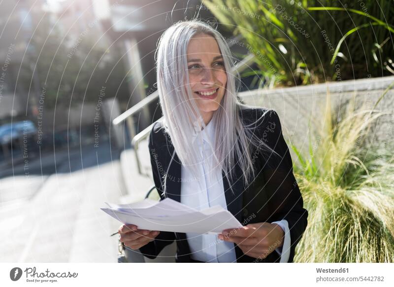 Lächelnde junge Geschäftsfrau sitzt mit Dokumenten auf einer Mauer in der Stadt sitzen sitzend Papiere Unterlagen Frau weiblich Frauen lächeln Geschäftsfrauen
