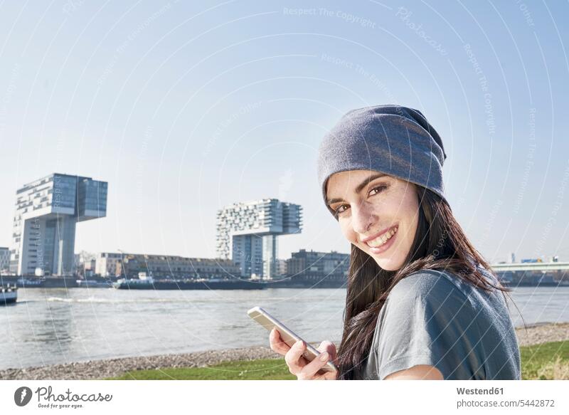 Deutschland, Köln, Porträt einer lächelnden jungen Frau mit Handy am Rhein weiblich Frauen Mobiltelefon Handies Handys Mobiltelefone Erwachsener erwachsen