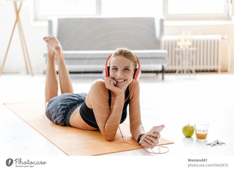 Lächelnde junge Frau in Sportkleidung liegt auf Gymnastikmatte und hört Musik liegen liegend Gymnastikmatten Matte Matten Sportbekleidung Sportdress Kleidung