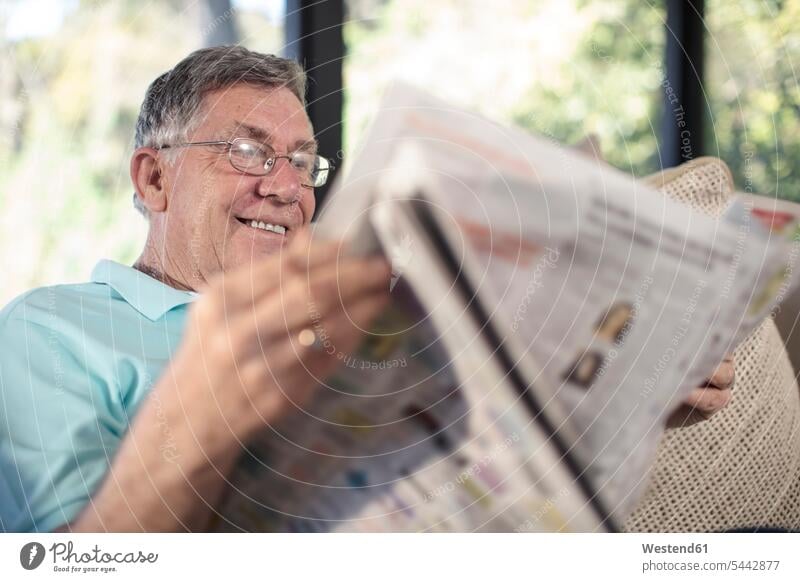 Lächelnder älterer Mann sitzt auf Couch und liest Zeitung Männer männlich Senior ältere Männer Senioren sitzen sitzend Zeitungen lesen Lektüre lächeln Sofa