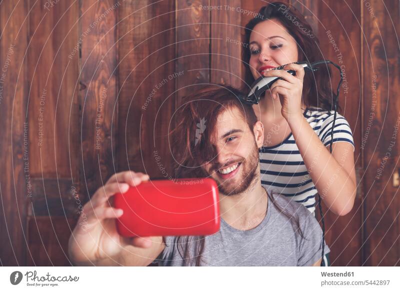 Junger Mann, der ein Selfie macht, während er von seiner Freundin mit einer Haarschneidemaschine einen Haarschnitt bekommt schneiden fotografieren Selfies Paar