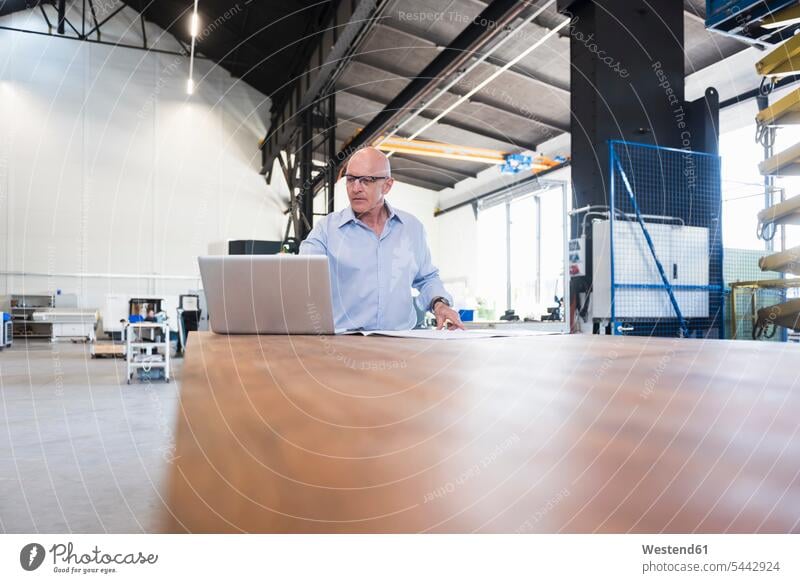 Mann mit Laptop schaut auf den Plan auf dem Tisch in der Fabrik Fabriken Notebook Laptops Notebooks Pläne Geschäftsmann Businessmann Businessmänner