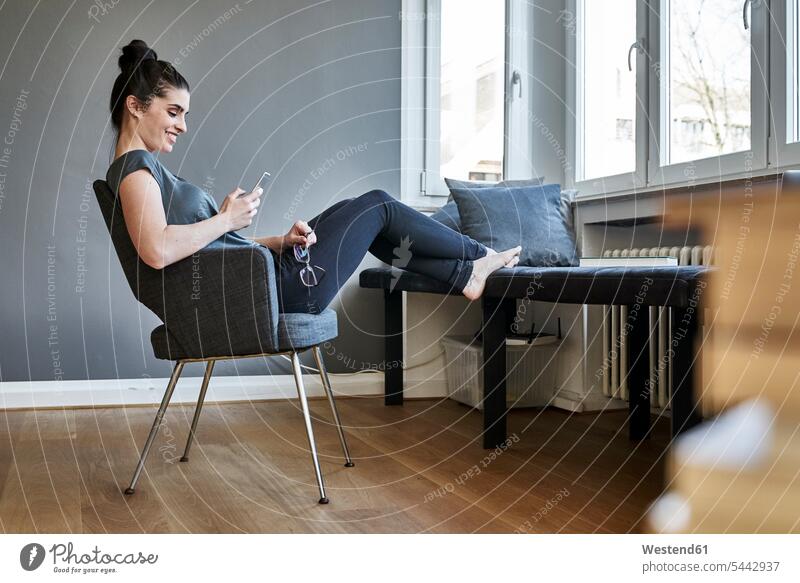 Lächelnde junge Frau benutzt Mobiltelefon zu Hause lächeln weiblich Frauen sitzen sitzend sitzt Handy Handies Handys Mobiltelefone Erwachsener erwachsen Mensch