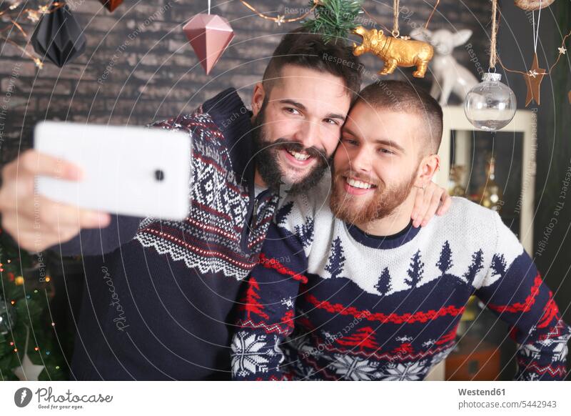 Porträt eines glücklichen schwulen Paares, das zu Weihnachten zu Hause ein Selfie mit dem Smartphone macht Schwules Paar Schwuler Portrait Porträts Portraits