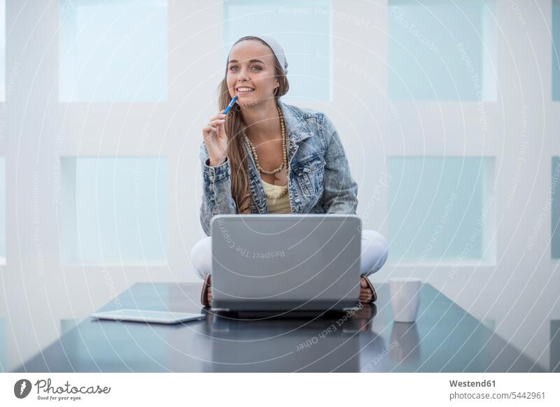 Junge Frau, die im Büro arbeitet und im Schneidersitz mit ihrem Laptop auf dem Schreibtisch sitzt Büroangestellte glücklich Glück glücklich sein glücklichsein