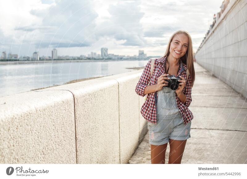 Lächelnde junge Frau mit einer Kamera am Flussufer Fotoapparat Fotokamera lächeln Portrait Porträts Portraits weiblich Frauen Fluesse Fluß Flüsse Erwachsener