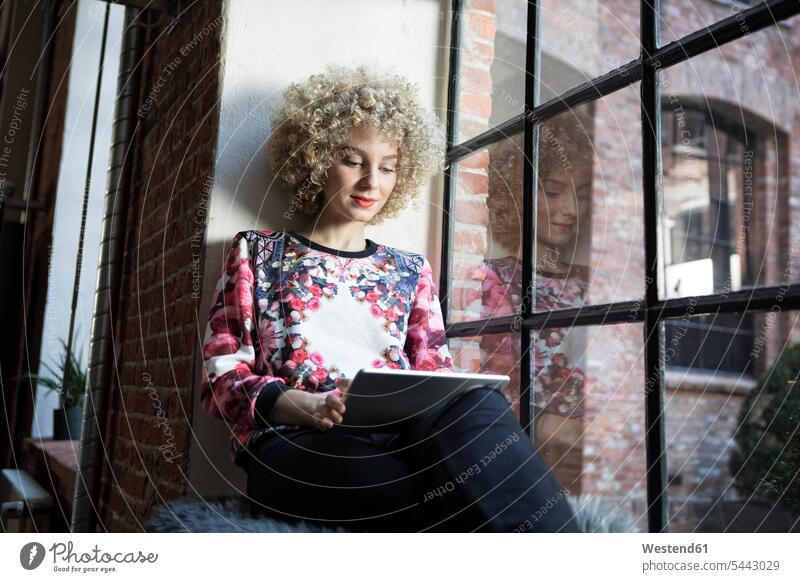 Junge Frau sitzt auf dem Fensterbrett und benutzt ein digitales Tablett weiblich Frauen Fensterbank Fenstersims Fensterbänke Zuhause zu Hause daheim lesen