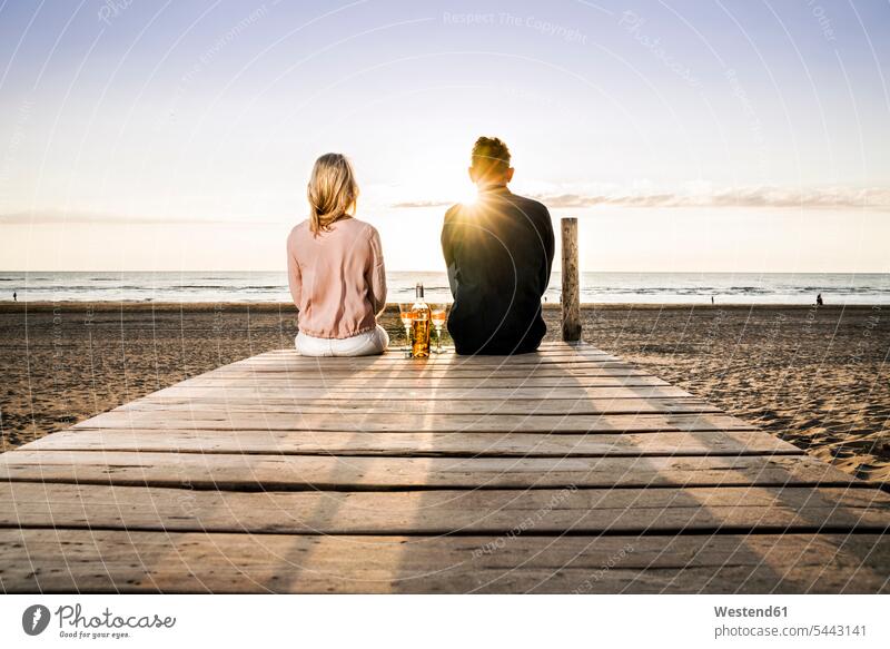 Paar mit Weingläsern, das bei Sonnenuntergang auf der Strandpromenade sitzt Beach Straende Strände Beaches entspannt entspanntheit relaxt sitzen sitzend Weine
