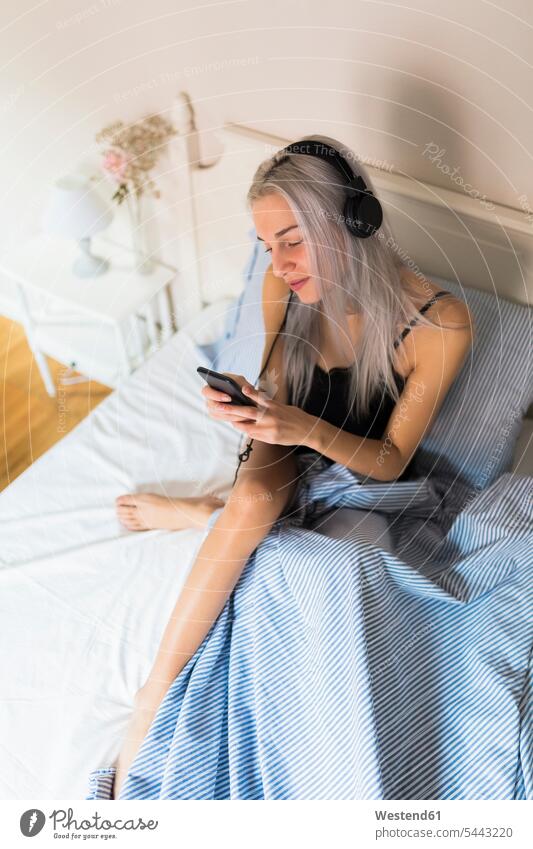 Junge Frau im Bett mit Handy und Kopfhörern Betten Kopfhoerer weiblich Frauen Mobiltelefon Handies Handys Mobiltelefone Erwachsener erwachsen Mensch Menschen