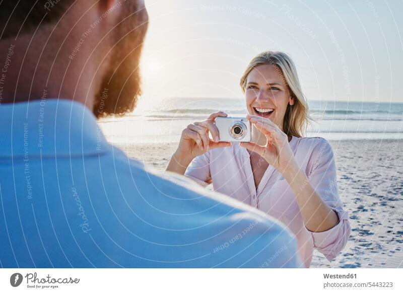 Glückliche Frau beim Fotografieren eines Mannes am Strand Beach Straende Strände Beaches fotografieren Paar Pärchen Paare Partnerschaft Fotoapparat Kamera