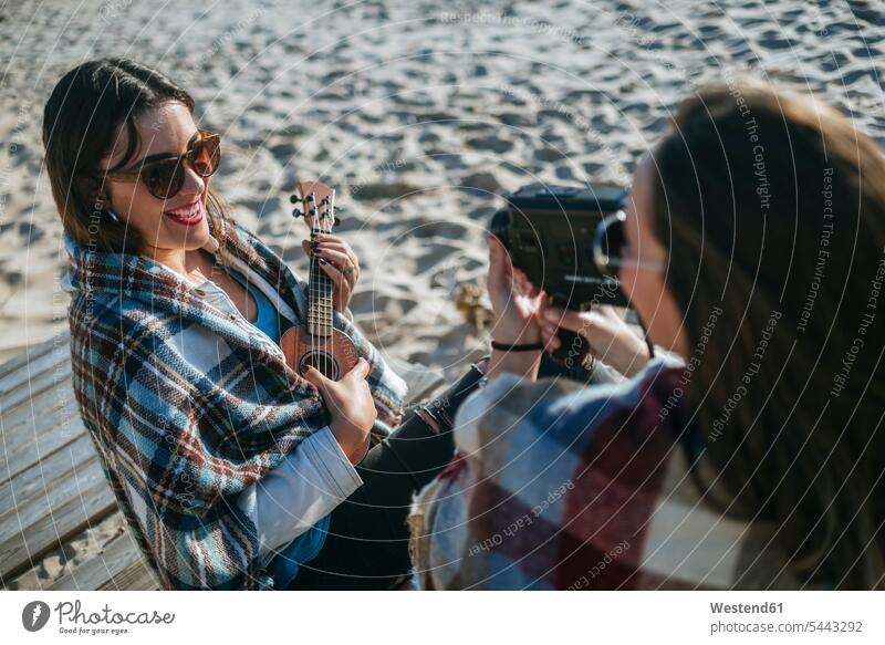 Lächelnde junge Frau spielt Ukulele, während ihr Freund mit Videokamera aufnimmt Freundinnen Strand Beach Straende Strände Beaches Freunde Freundschaft