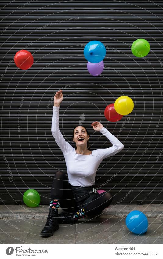 Lachende junge Frau sitzt auf dem Bürgersteig und beobachtet fliegende Luftballons weiblich Frauen Ballons Luftballone lachen Erwachsener erwachsen Mensch